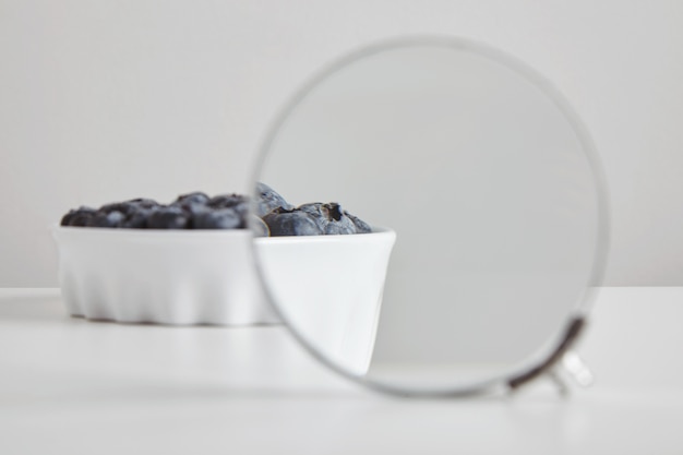 자세한 내용을 보려면 쌍안경 돋보기를 통해 확대 흰색 테이블에 고립 된 건강한 식습관과 영양을위한 세라믹 그릇 개념에 블루 베리 항산화 유기농 슈퍼 푸드의 힙