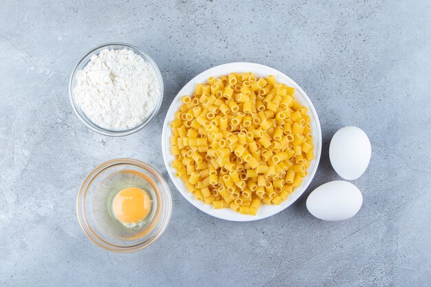 Heap of raw pipette rigate pasta in a white bowl with eggs and flour. 