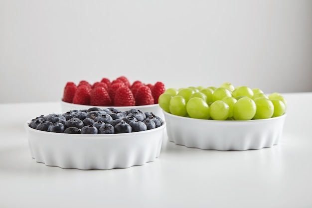 Heap of ripe raspberries and blueberries and green seedless muscat grapes accurately placed in ceramic bowls isolated on white table