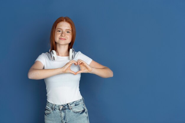 Heart gesture. Caucasian young girl's portrait on blue wall. Beautiful female redhair model with cute freckles. Concept of human emotions, facial expression.