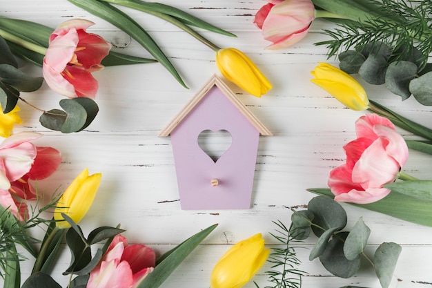 Heart shape bird house surrounded with pink and yellow tulips on white wooden desk