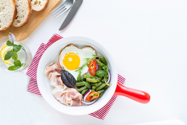 Heart shaped egg, bacon, green beans on a sauce pan, bread on cutting board and water with lemon