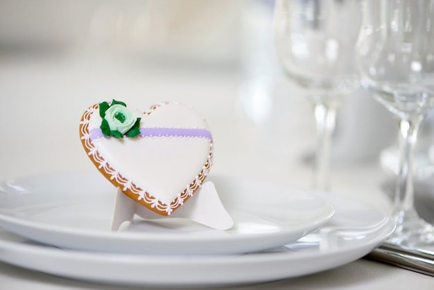 Free photo heart shaped glazed cookie - decorated with green glaze flower and tiny pattern stands on a white plates as a decoration for festive wedding table near the wine glasses