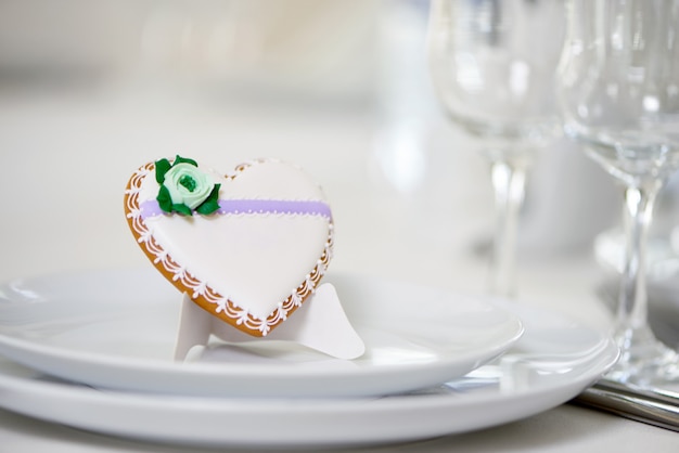 하트 모양의 유약 쿠키-녹색 유약 꽃과 작은 패턴으로 장식되어 와인 잔 근처의 축제 웨딩 테이블 장식으로 흰색 접시에 서 있습니다.