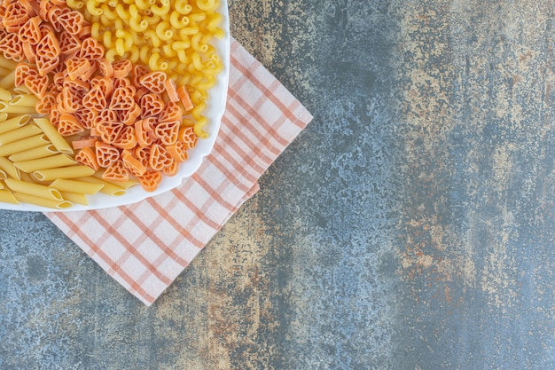 Heart shaped, penne and fusilli pastas in bowl on towel, on the marble background.