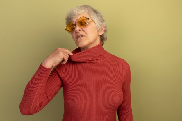 Heated old woman wearing red turtleneck sweater and sunglasses pulling collar of her sweater with closed eyes isolated on olive green wall with copy space