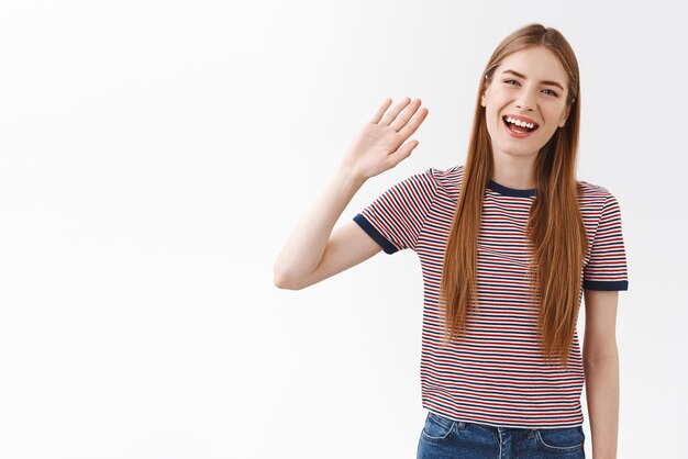 Hello how are you Cheerful positive goodlooking girl student in striped tshirt raise hand and waving friendly smiling toothy saying hi greeting you gladly standing white background upbeat