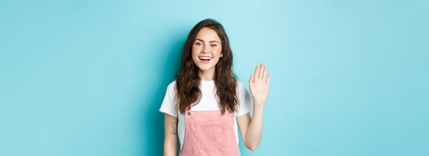 Free photo hello nice to meet you beautiful friendly girl waving hand to say hi looking happy and smiling while greeting you giving warm welcome standing over blue background