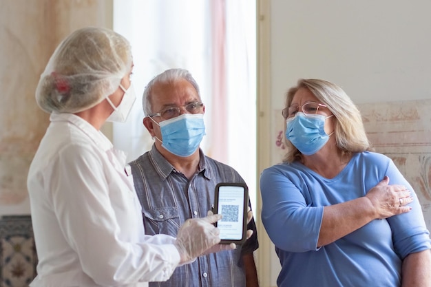 Helpful nurse explaining patients how use QR-code. Doctor in uniform holding mobile phone, showing QR-code to elderly couple. Medicine, vaccination, immunization, healthcare concept