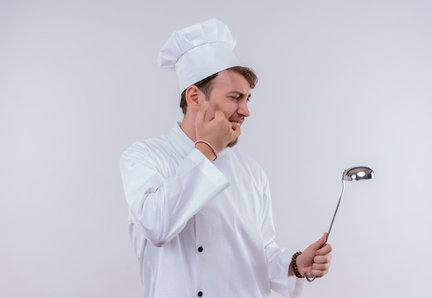 A helpless young bearded chef man in white uniform burning his hand while touching hot ladle on a white wall