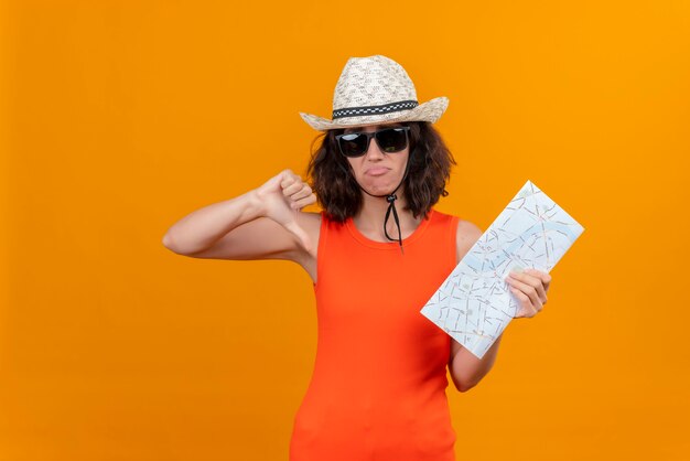親指を下に示す地図を保持している太陽の帽子とサングラスを身に着けているオレンジ色のシャツの短い髪の無力な若い女性
