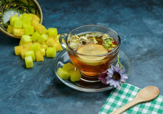 Herbal lemony tea with sugar cubes, spoon, tea towel in a cup on dark blue surface, high angle view.