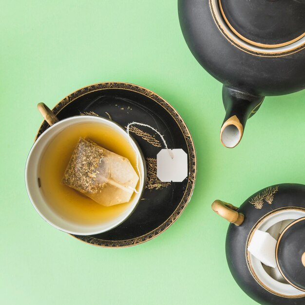 Herbal tea bag in the cup with teapot and sugar cubes on green background