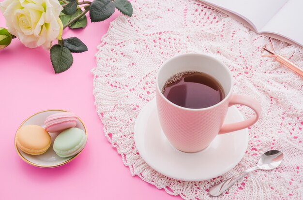 Herbal tea cup with macaroons; spoon; rose; pen and book on pink background