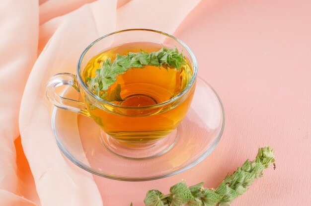 Herbal tea with herbs in a glass cup on pink and textile, high angle view.