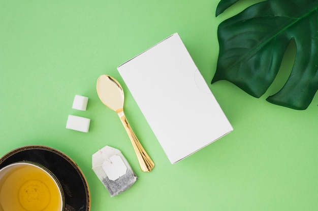 Herbal tea with sugar cubes, tea bag, spoon and box on green background