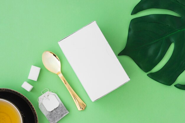 Herbal tea with sugar cubes, tea bag, spoon, leaf and box on green background