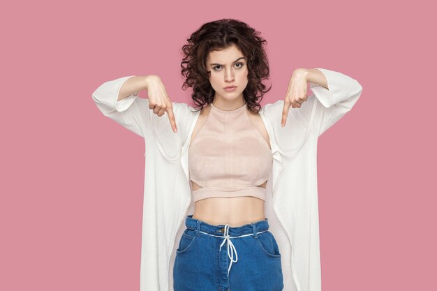 Here and right now. portrait of serious cute brunette young woman with curly hairstyle in casual style standing and pointing down and looking at camera. indoor studio shot isolated on pink background.