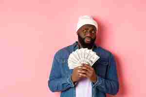 Free photo hesitant african-american man holding money, looking left with doubts and concerns, standing against pink background.