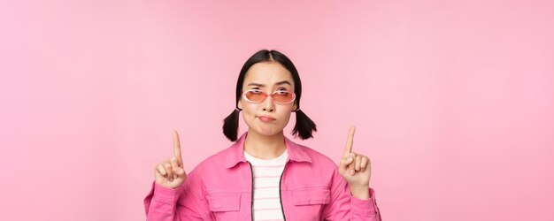 Hesitant asian girl wears sunglasses frowns and points fingers up looks with disbelief skeptical face expression stands over pink background