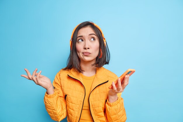  hesitant Asian woman spreads palms looks above with clueless expression dressed in orange jacket uses smartphone listens music from playlist via wireless headphones isolated on blue