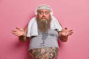 Free photo hesitant bearded adult man spreads palms and looks confused, has regular workout for weight loss, listens music in headphones, wears headband and undersized t shirt, tattooed belly sticking out