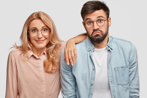 Hesitant bearded man stands near his blonde woman colleague who has satisfied expression