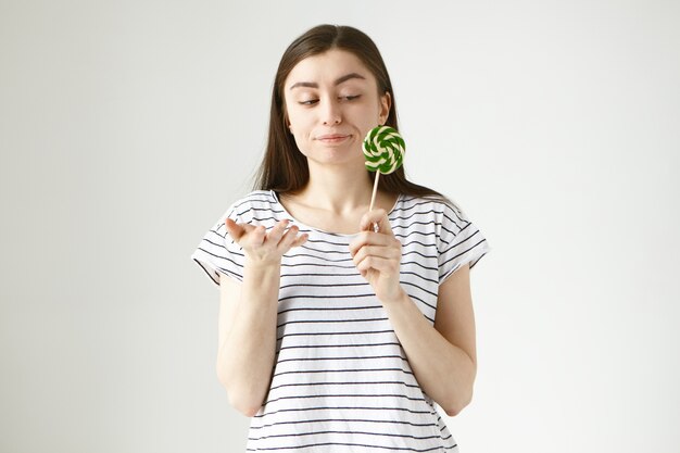 hesitant brunette woman in striped t-shirt making uncertain gesture, holding spiral round hard candy in her hand and pursing lips