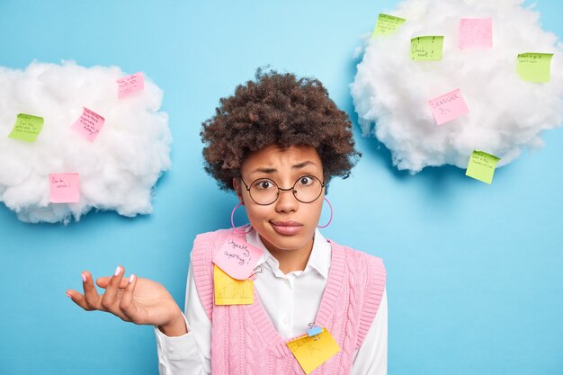 Hesitant clueless Afro American schoolgirl poses around paper stickers with written tasks to do uses post it for new ideas wears round spectacles white shirt pink vest