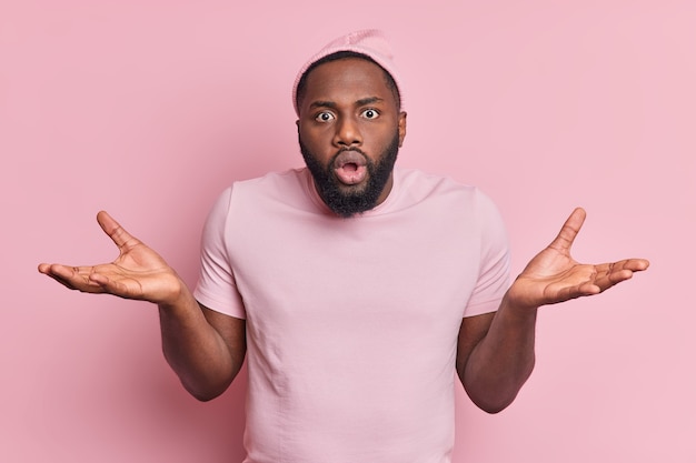 Free photo hesitant confused shocked black man spreads palms puzzled by difficult situation looks surprised and unaware wears hat t shirt isolated over pink wall