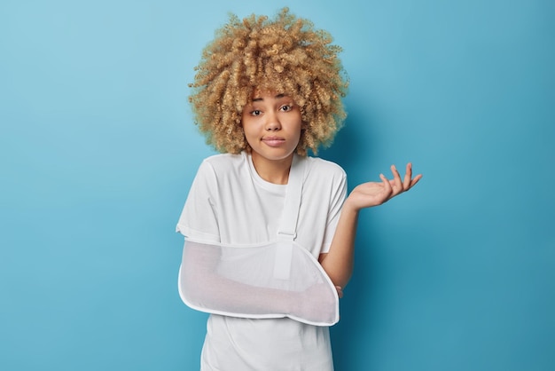 Hesitant curly haired woman has broken arm wears soft splint armband shrugs shoulders feels unaware or questioned dressed in white t shirt isolated over blue background Personal accident concept