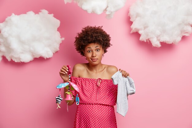 Hesitant curly haired woman holds two items and necessities for baby, holds childs clothes and mobile, thinks what better to buy for newborn, has big pregnant belly. Pregnancy and birth concept