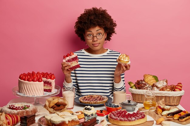 Hesitant dark skinned curly woman feels doubt which piece of cake to choose, has temptation to eat junk food, poses at big festive table with desserts against pink background