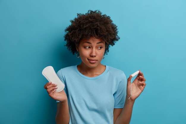 주저하는 어두운 피부를 가진 여성은 사용하지 않는 깨끗한 생리대와 탐폰을 들고 월경에 가장 친밀한 제품을 선택하고 건강에 관심이 있으며 파란색 벽에 격리됩니다. 여성, 위생, 산부인과 개념
