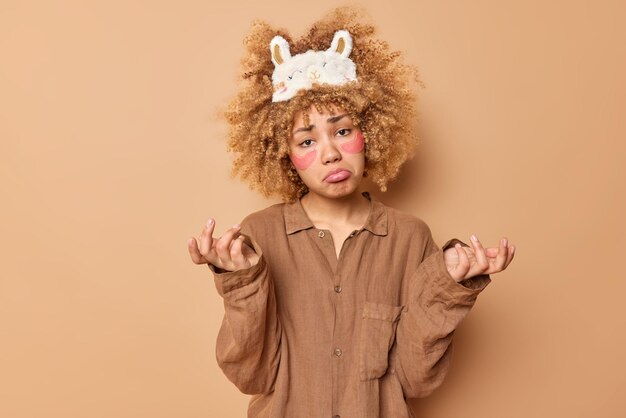 Hesitant displeased woman with curly bushy blonde hair applies beauty patches shrugs shoulders has sulking expression dressed in slumber suit doesnt know what to do isolated over brown background