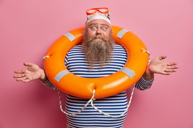 Hesitant doubtful bearded man spreads hands sideways, feels confused, wears swimming hat, goggles and sailor t shirt, poses with inflated lifebuouy isolated on pink wall. Overweight lifesaver at beach