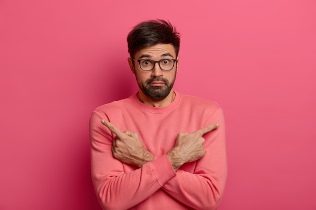 Free photo hesitant embarrassed bearded man points sideways, keeps arms crossed over body, picks necessary object, has doubtful surprised expression, dressed in bright pink jumper. two options or variants