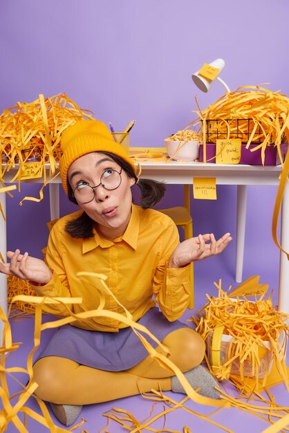 Hesitant female model in yellow hat shirt and skirt poses crossed legs near desktop spreads palms has no clue or idea what to do first sits on floor in own cabinet surrounded py paper wastes.