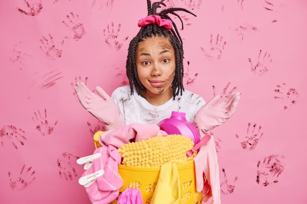 Hesitant housewife spreads palms and looks confused poses near laundry basket being dirty after washing poses against pink wall