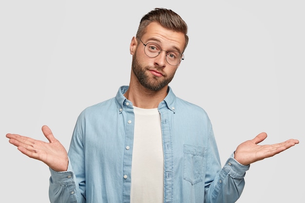 Hesitant puzzled unshaven man shruggs shoulders in bewilderment, feels indecisive, has bristle, trendy haircut, dressed in blue stylish shirt, isolated on white wall. Clueless male poses indoor