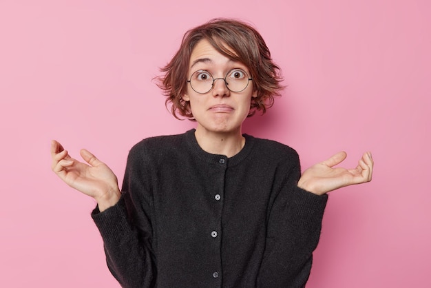 Hesitant puzzled young European woman with short hairstyle spreads palms looks clueless cannot mae decision wears round spectacles and black jumper isolated over pink background. So what to do