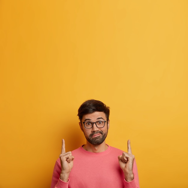 Hesitant surprised man points upwards with confused expression, points on blank space, has doubts and asks your opinion, makes perplexed choice, wears spectacles and casual jumper, yellow wall