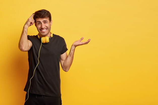 Hesitant unaware man scratches head, makes difficult decision, raises palm with hesitation, wears casual black t shirt, headphones on neck for listening music