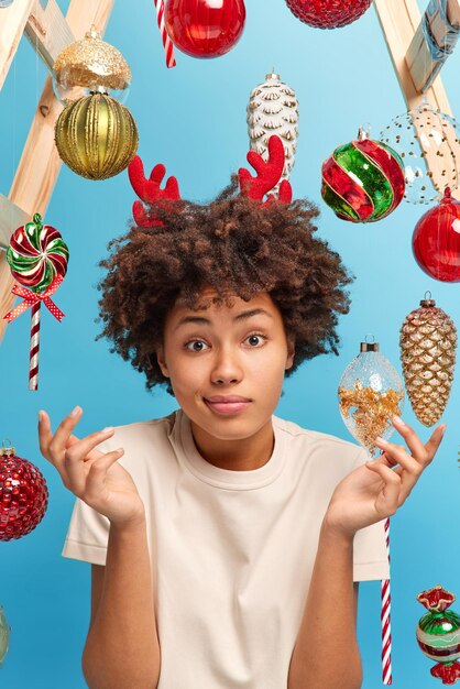 Hesitant woman shrugs shoulders feels doubtful cannot understand something dressed in casual t shirt poses against blue background with new year toys for decoration. I wonder how to decorate fir tree
