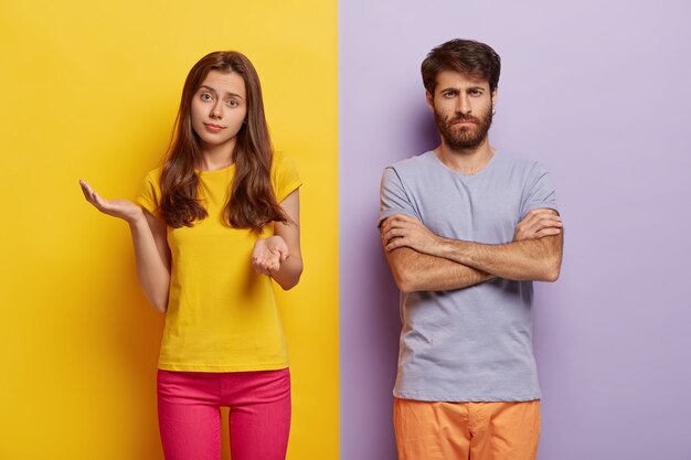 Hesitant young woman spreads hands with doubt, wears casual clothes, dissatisfied man keeps hands crossed, discontent with something