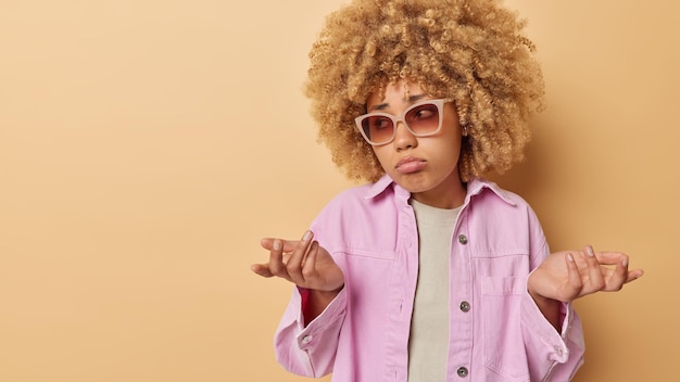 Free photo hesitant young woman with curly hair shrugs shoulders in bewilderment feels doubt feels uncertain wears sunglasses and pink jacket isolated over beige background with empty space for promotion