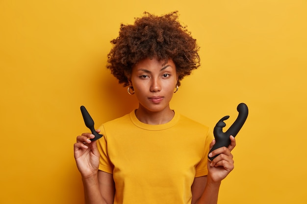 躊躇しているアフリカ系アメリカ人の女性は、黄色のTシャツを着た、貫通プレイの前にウォームアップするためのバットプラグ、膣刺激用のウサギの形をしたバイブレーターを持っています。屋内で大人のおもちゃを持つ若い女性