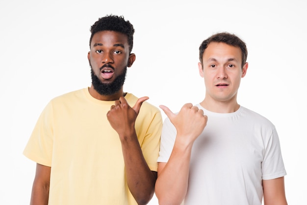 Hey bro. Two young handsome men standing on white isolated wall and pointing on each other with index fingers