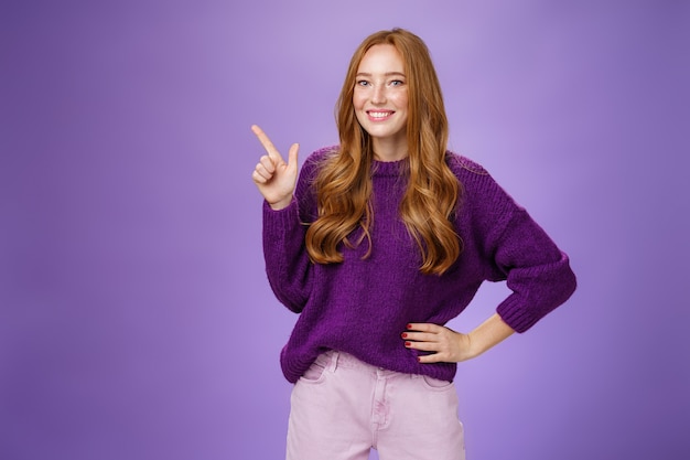 Hey click here. Portrait of friendly-looking charming and cute redhead female in purple sweater pointing at upper left corner with index finger and smiling, showing copy space, recommending, offering.