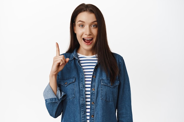 Hey got excellent idea. Excited young woman has suggestion, pointing finger up, smiling and telling big news, advertising smth above head, standing against white background.